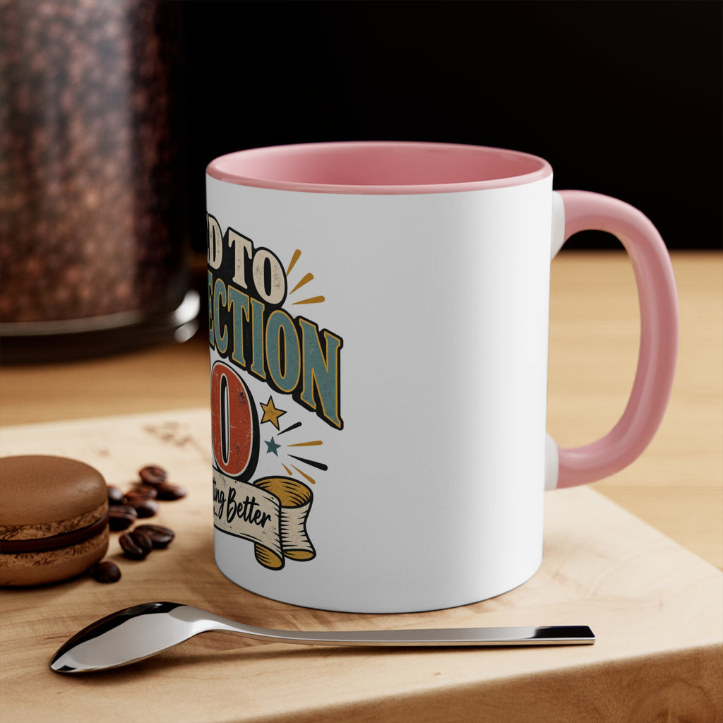 A white ceramic coffee mug with a pink handle and interior, featuring a retro-style graphic that reads 'aged to perfection.' The mug sits on a wooden cutting board with a spoon, coffee beans, and a macaron nearby, with a dark background.