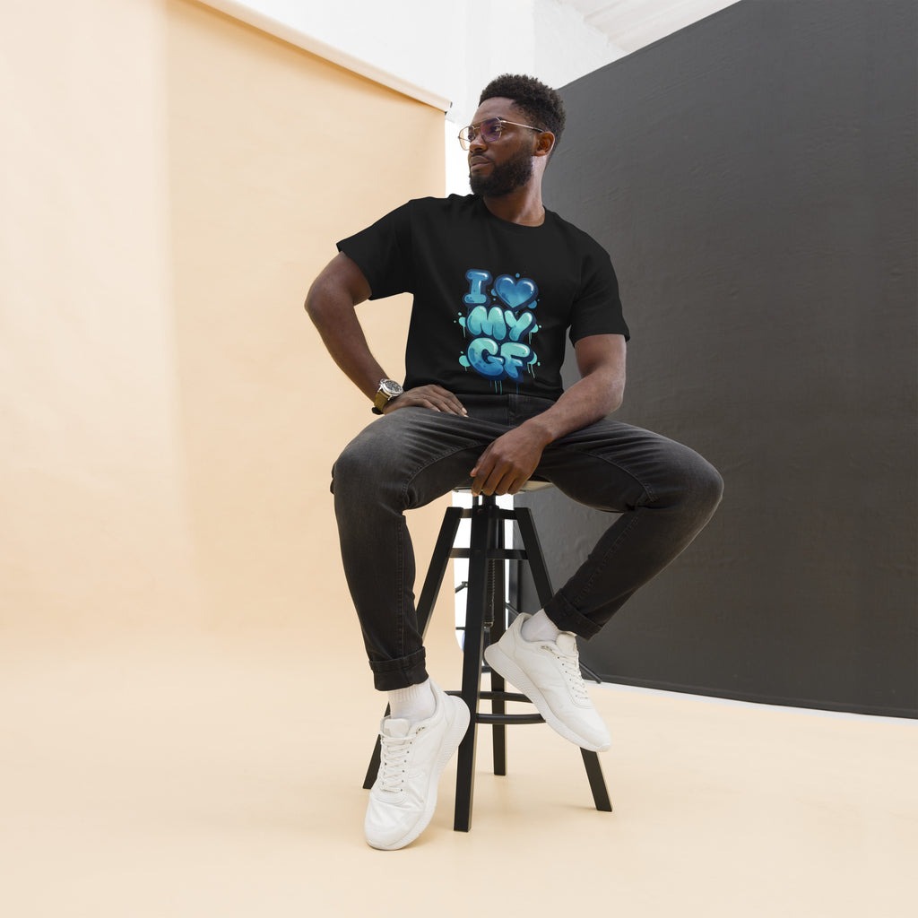 Man sitting on a stool wearing a green t-shirt with text, black pants, and white sneakers.