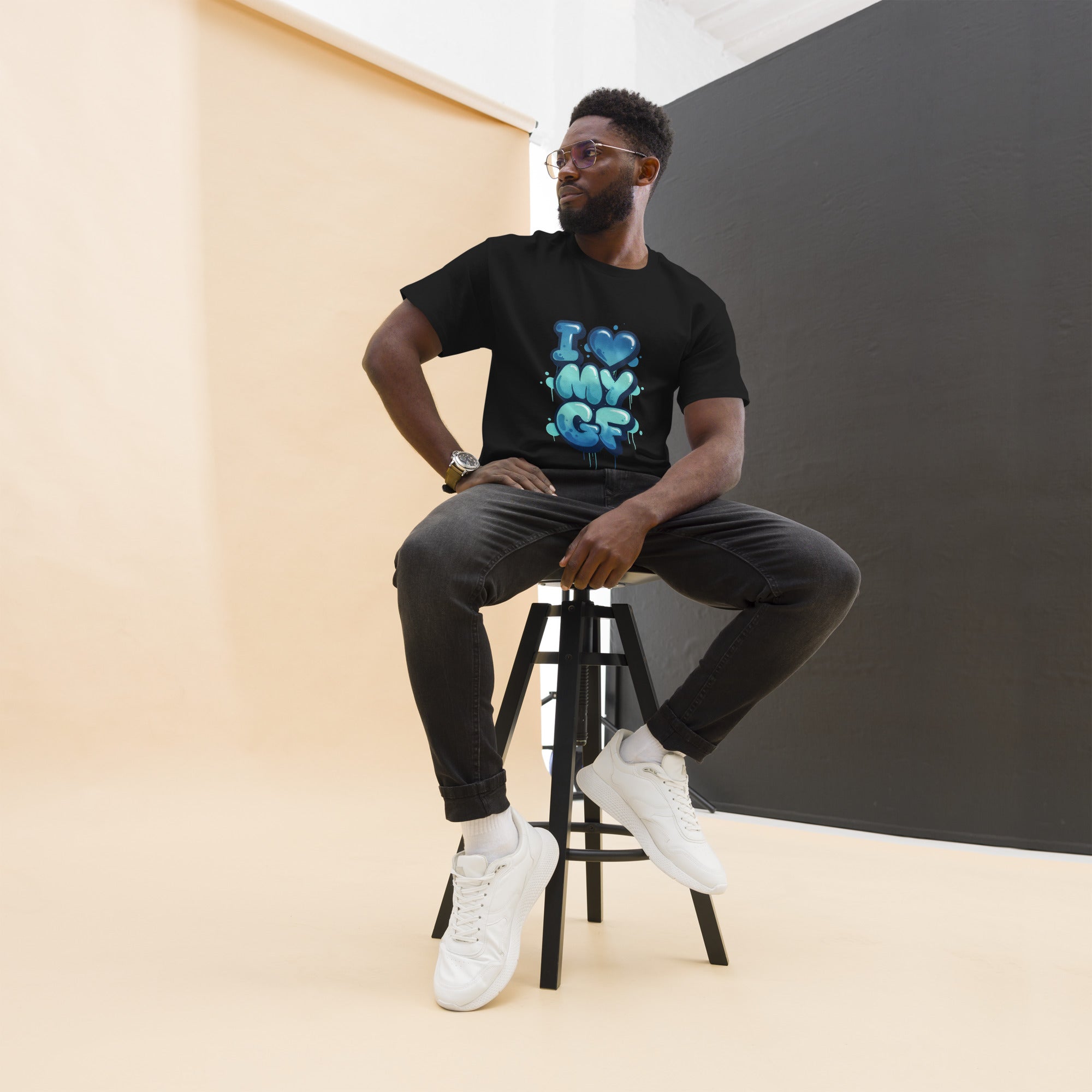 Man sitting on a stool wearing a green t-shirt with text, black pants, and white sneakers.