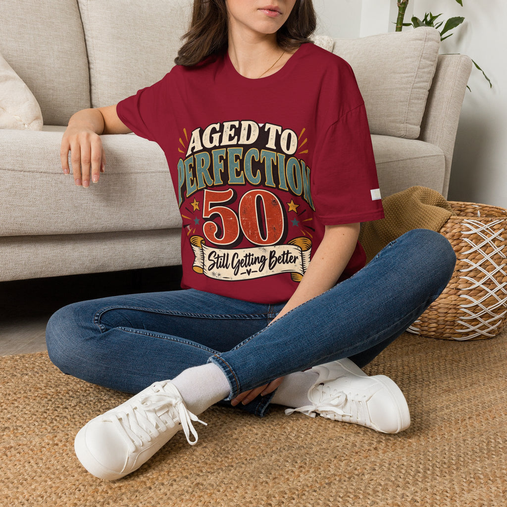 A woman sitting on a woven rug in front of a beige sofa, wearing a maroon graphic t-shirt with the text 'AGED TO PERFECTION 50 Still Getting Better' in green, orange, and white.