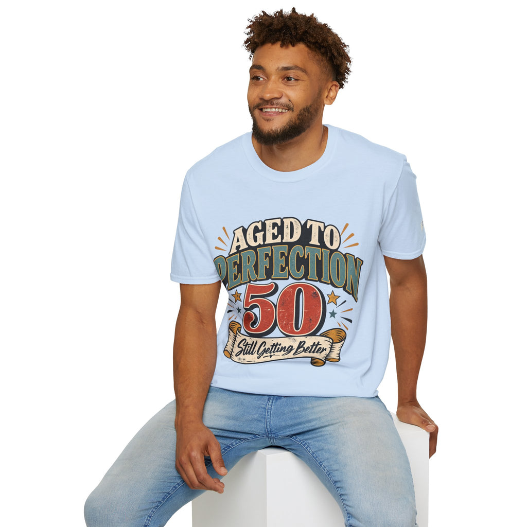A man wearing a light blue graphic t-shirt with a '50 Aged to Perfection Still Outfitting Better' print, paired with light blue jeans, sitting on a white block against a white background.