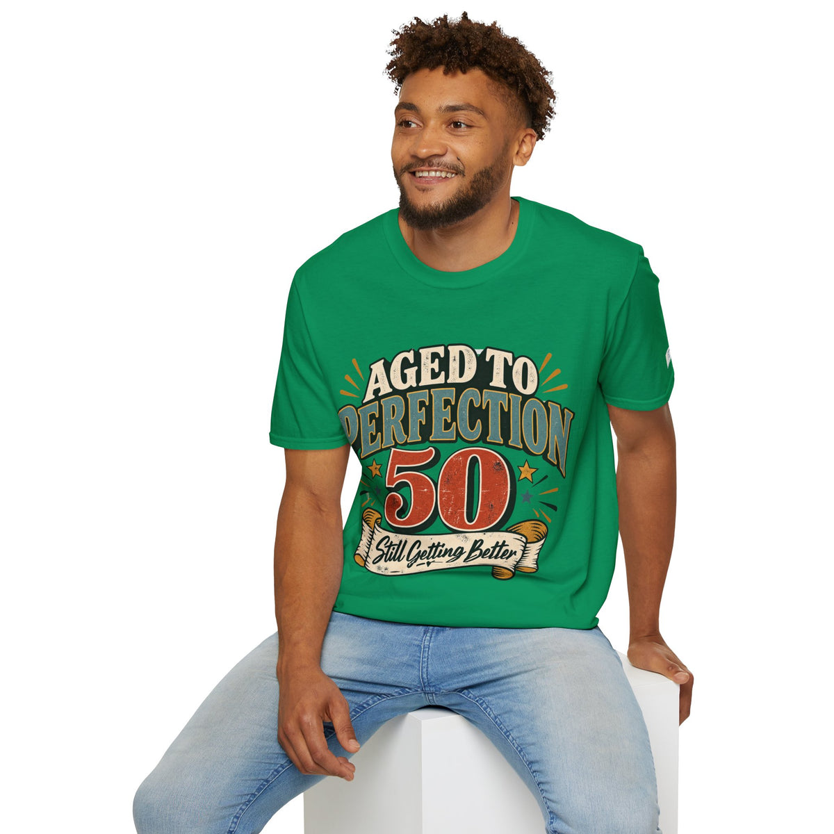 A man wearing a bright green graphic t-shirt with the text 'AGED TO PERFECTION 50 Still Outfit Better' and light blue jeans, sitting on a white block against a white background.
