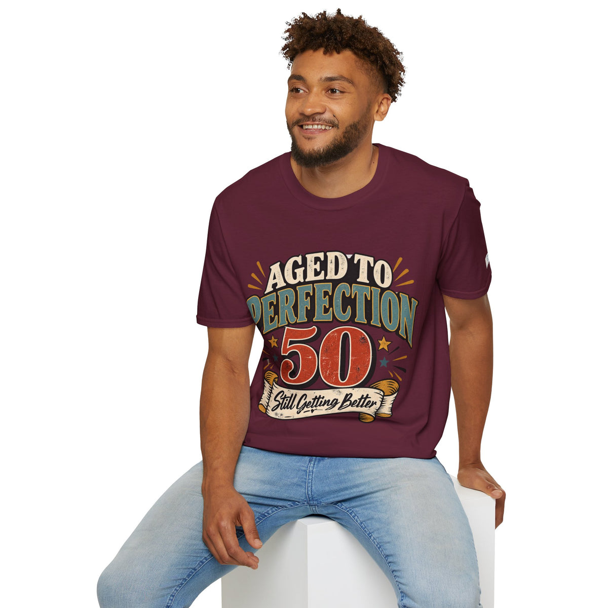 A man wearing a maroon graphic t-shirt with a retro-style 'Aged to Perfection 50' print, paired with light blue jeans, sitting on a white block against a plain white background.