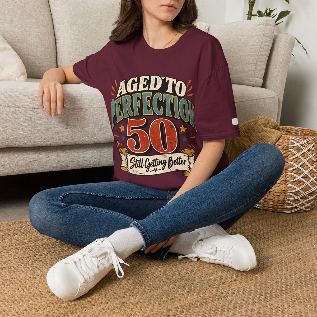 A woman sitting on a woven rug in front of a beige sofa, wearing a maroon graphic t-shirt with the text 'AGED TO PERFECTION 50 Still Getting Better' in distressed retro lettering, paired with blue jeans and white sneakers.