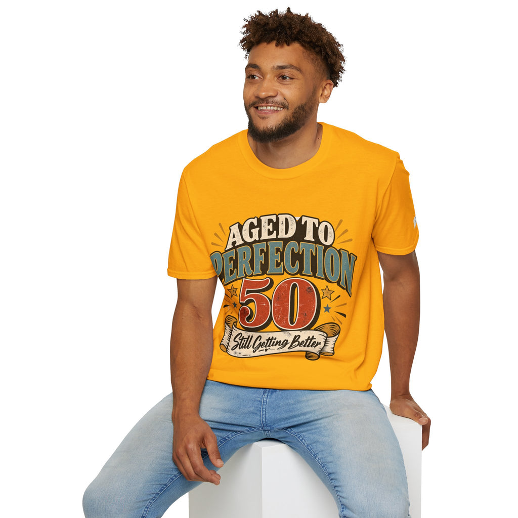 A man wearing a bright yellow graphic t-shirt with the text 'AGED TO PERFECTION 50 Still Outfit Better' and light blue jeans, sitting on a white block against a white background.