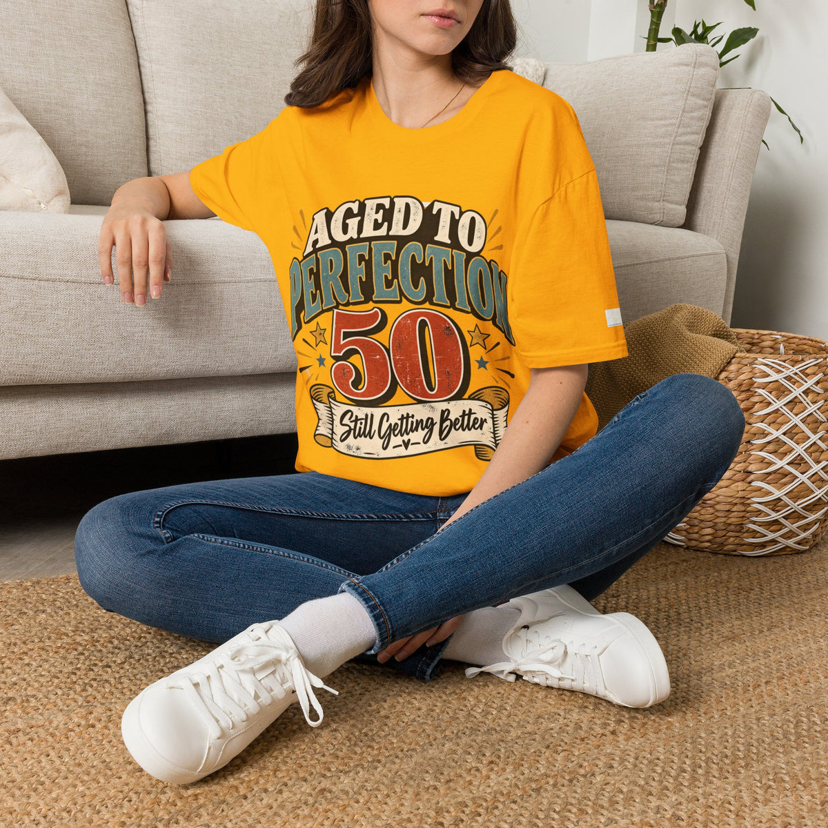A woman sits cross-legged on a woven rug in front of a beige sofa, wearing an oversized bright yellow graphic T-shirt with the text 'AGED TO PERFECTION 50 Still Getting Better' and blue jeans with white sneakers.
