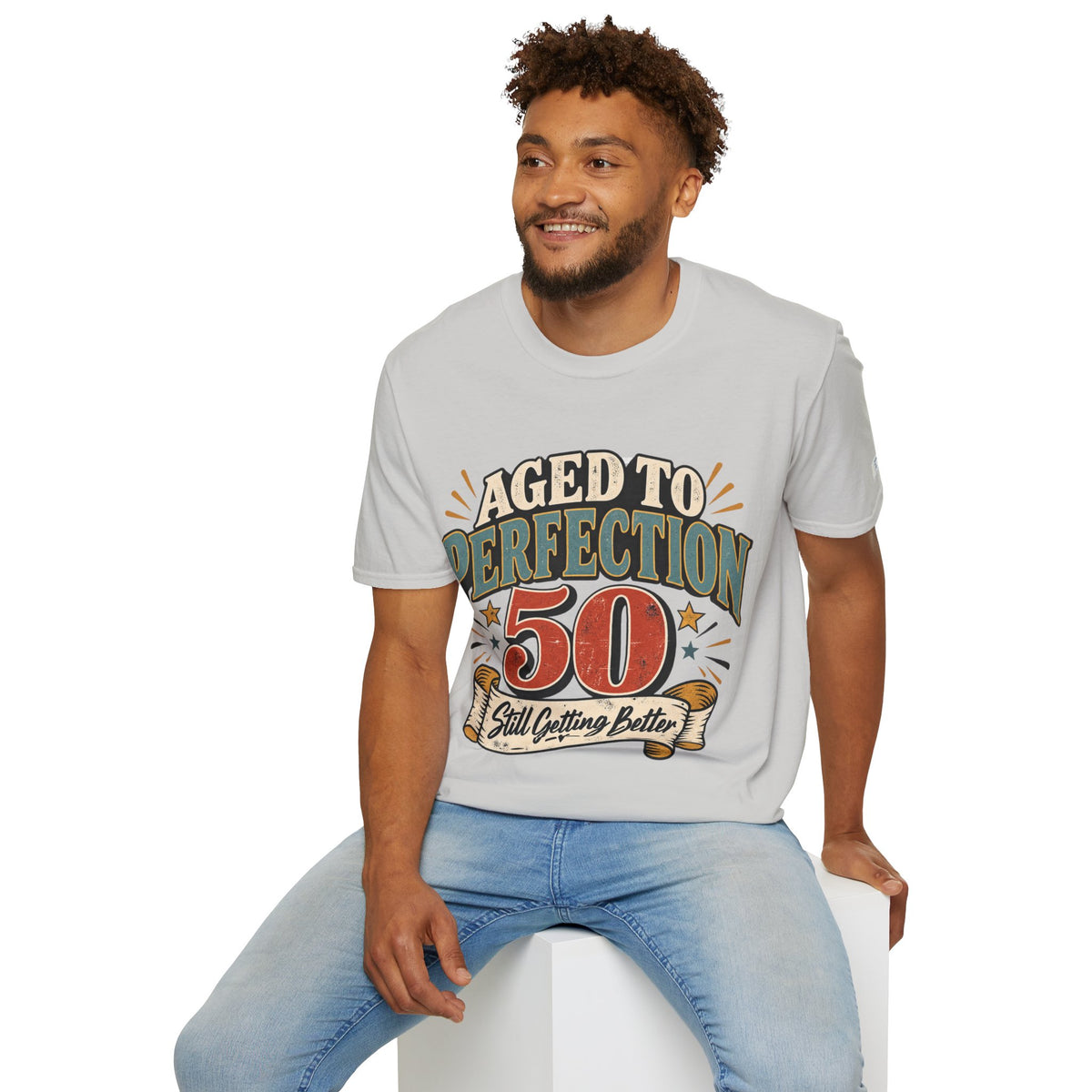 A man wearing a light gray graphic t-shirt with a retro-style 'Aged to Perfection 50 Still Outfitting Better' print, paired with light blue jeans, sitting on a white block against a white background.