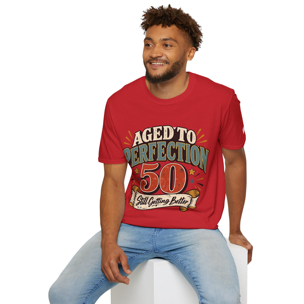 A man wearing a red graphic t-shirt with the text 'AGED TO PERFECTION 50 Still Outfit Better' and light blue jeans, sitting on a white block against a white background.