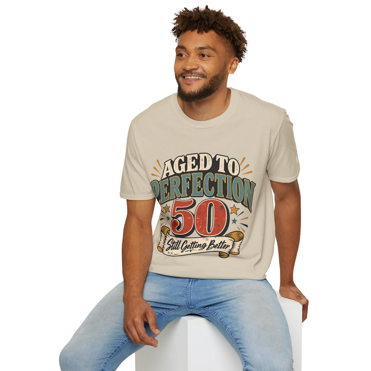 A man wearing a beige graphic t-shirt with a 'Aged to Perfection 50' print, sitting on a white block against a white background.