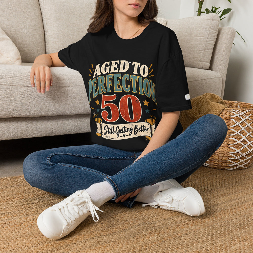 A woman sitting cross-legged on a woven rug in front of a beige sofa, wearing a black graphic T-shirt with the text 'AGED TO PERFECTION 50 Still Getting Better' in distressed multicolor lettering, paired with blue jeans and white sneakers.