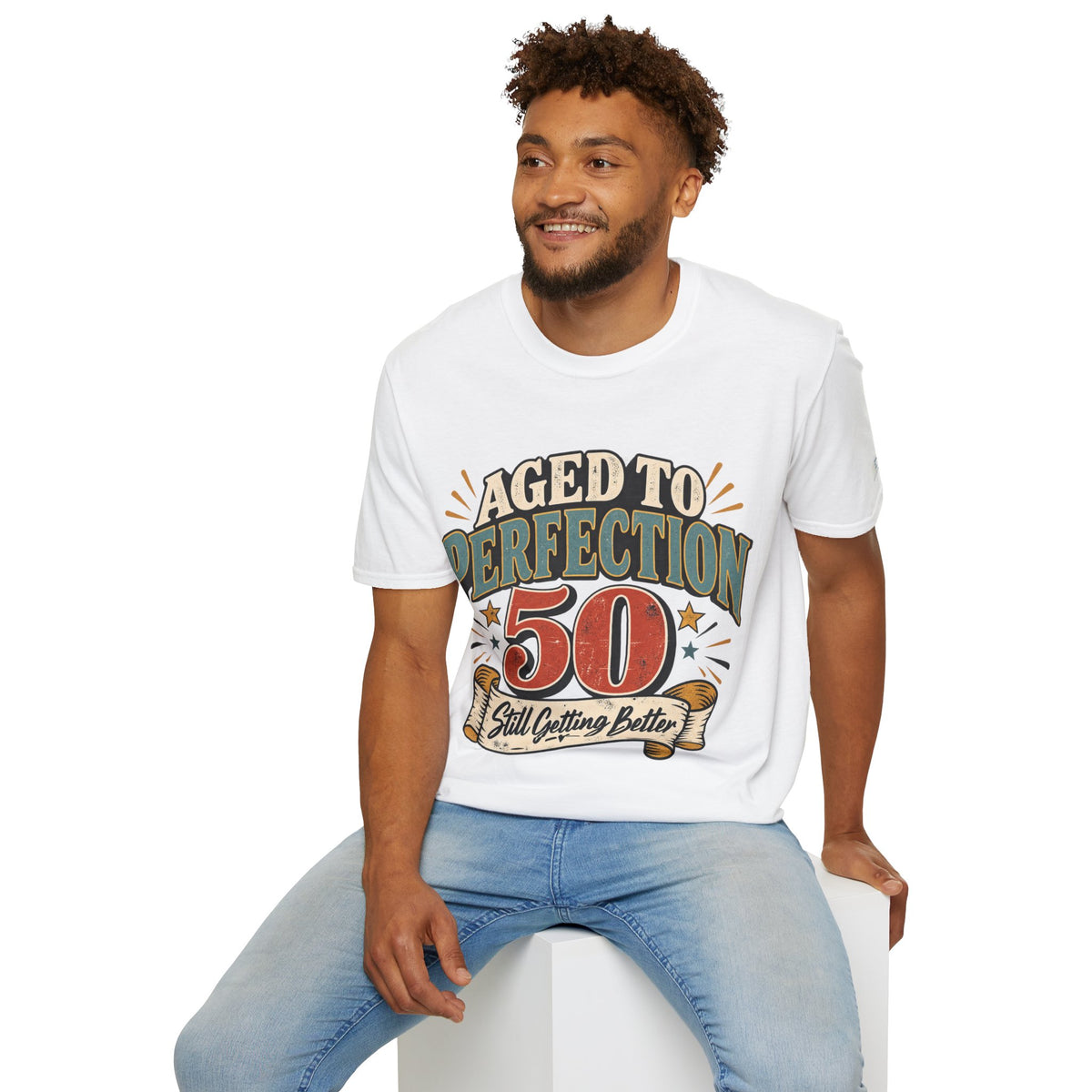 A man wearing a white graphic t-shirt with a vintage-style 'Aged to Perfection 50 Still Getting Better' print, paired with light blue jeans, sitting on a white block against a white background.