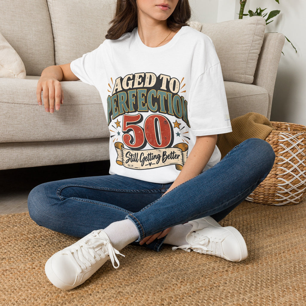 A woman sitting cross-legged on a woven rug in front of a beige sofa, wearing a white graphic T-shirt with the text 'AGED TO PERFECTION 50 Still Getting Better' in a retro multicolor design, paired with blue jeans and white sneakers.