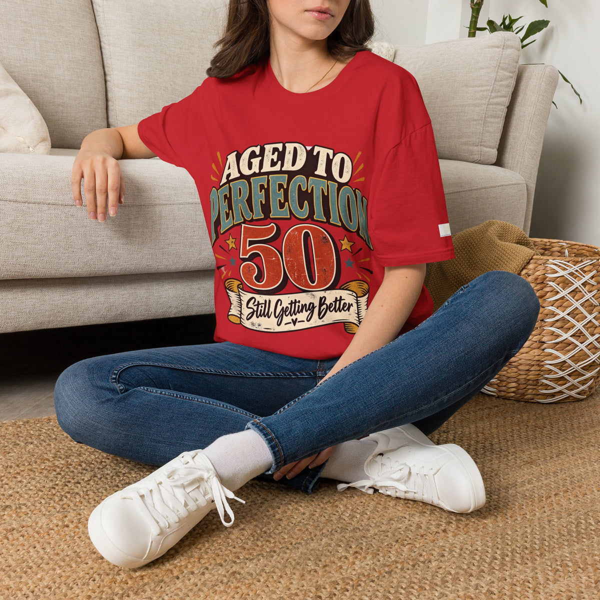 A woman sitting cross-legged on a woven rug in front of a beige sofa, wearing a red graphic T-shirt with the text 'AGED TO PERFECTION 50 Still Getting Better' and blue jeans with white sneakers.