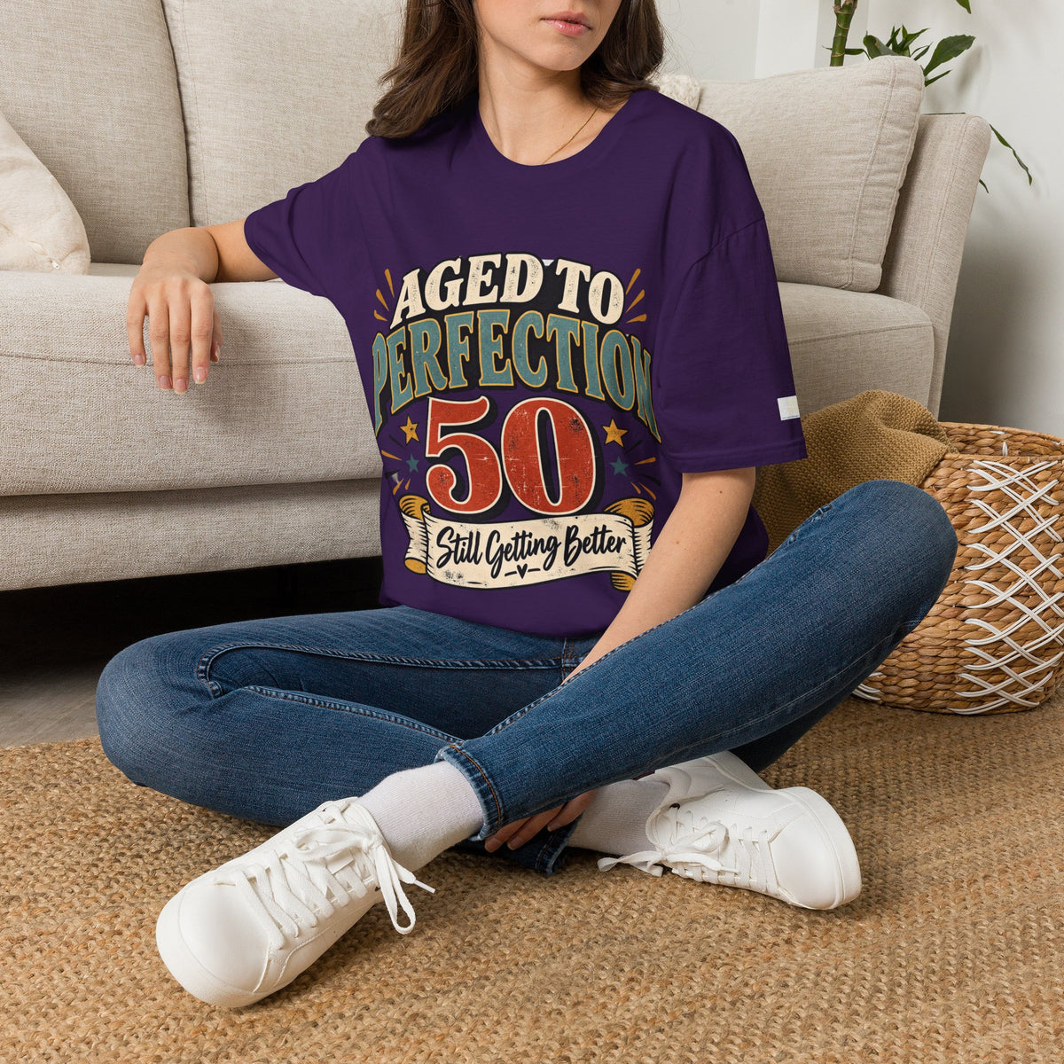 A woman sitting on a woven rug in front of a beige sofa, wearing a purple graphic t-shirt with the text 'AGED TO PERFECTION 50 Still Getting Better' in a retro design.