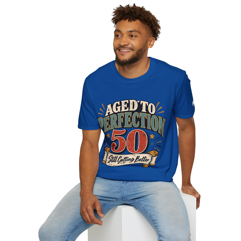 A man wearing a royal blue graphic t-shirt with the text 'AGED TO PERFECTION 50 Still Outing Better' in a retro design, paired with light blue jeans, sitting on a white block against a white background.