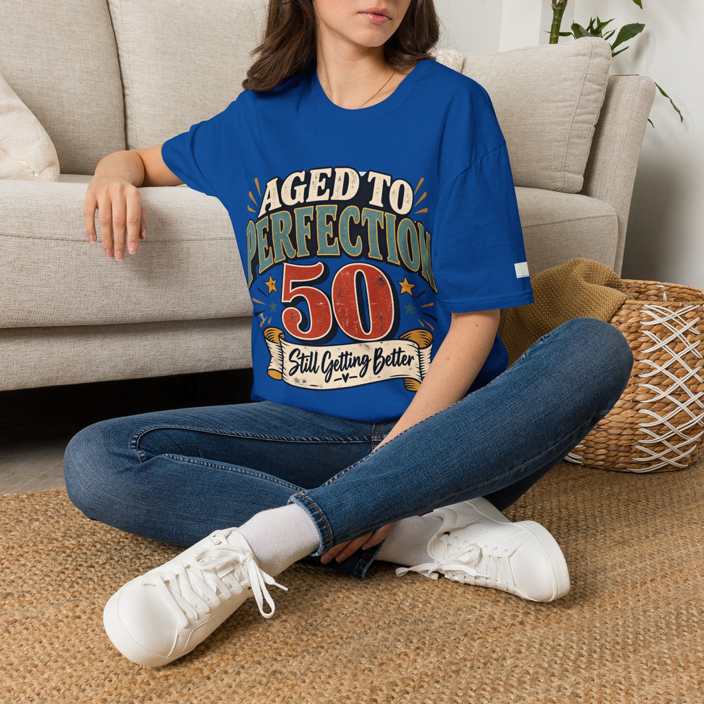 A woman sitting cross-legged on a woven rug in front of a beige sofa, wearing a royal blue graphic T-shirt that reads 'TEACHERS PLANT THAT GROW FOREVER' with an apple, books, and leaves design, paired with blue jeans and white sneakers.