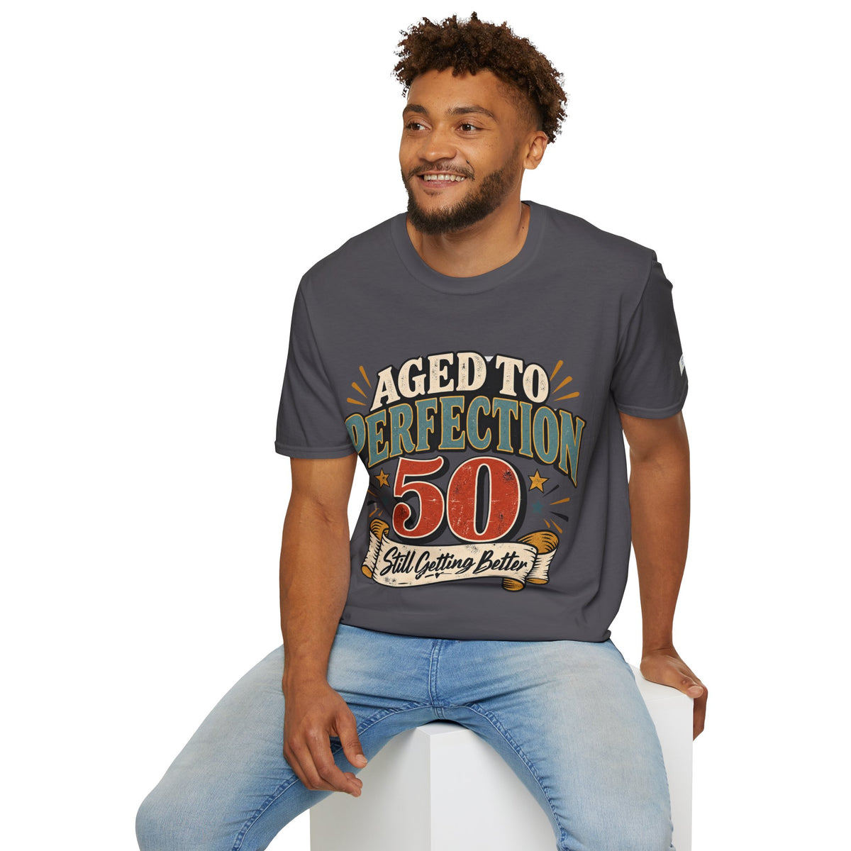 A man wearing a dark gray graphic t-shirt with a retro-style 'Aged to Perfection 50' print, paired with light blue jeans, sitting on a white block against a plain white background.
