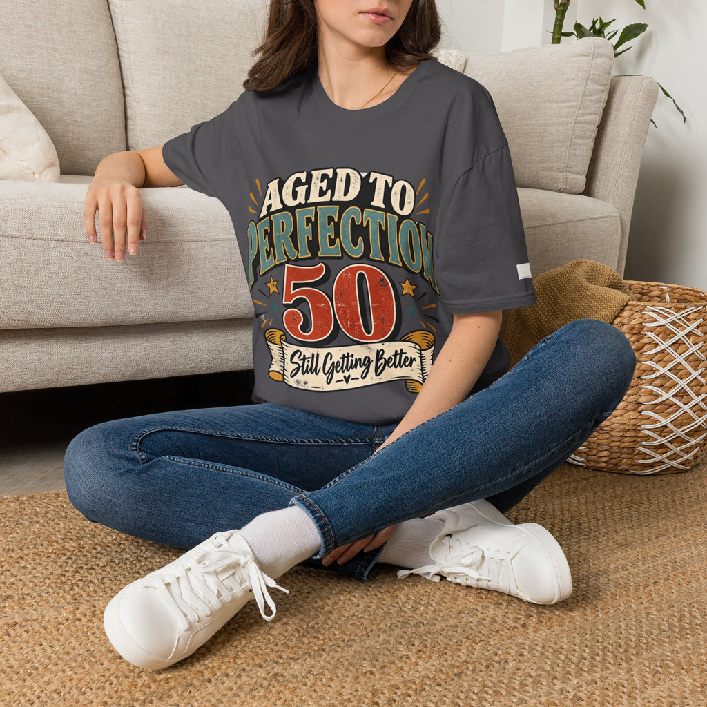 A woman sitting cross-legged on a woven rug in front of a beige sofa, wearing a dark gray graphic t-shirt with a moose and campfire design, blue jeans, and white sneakers.