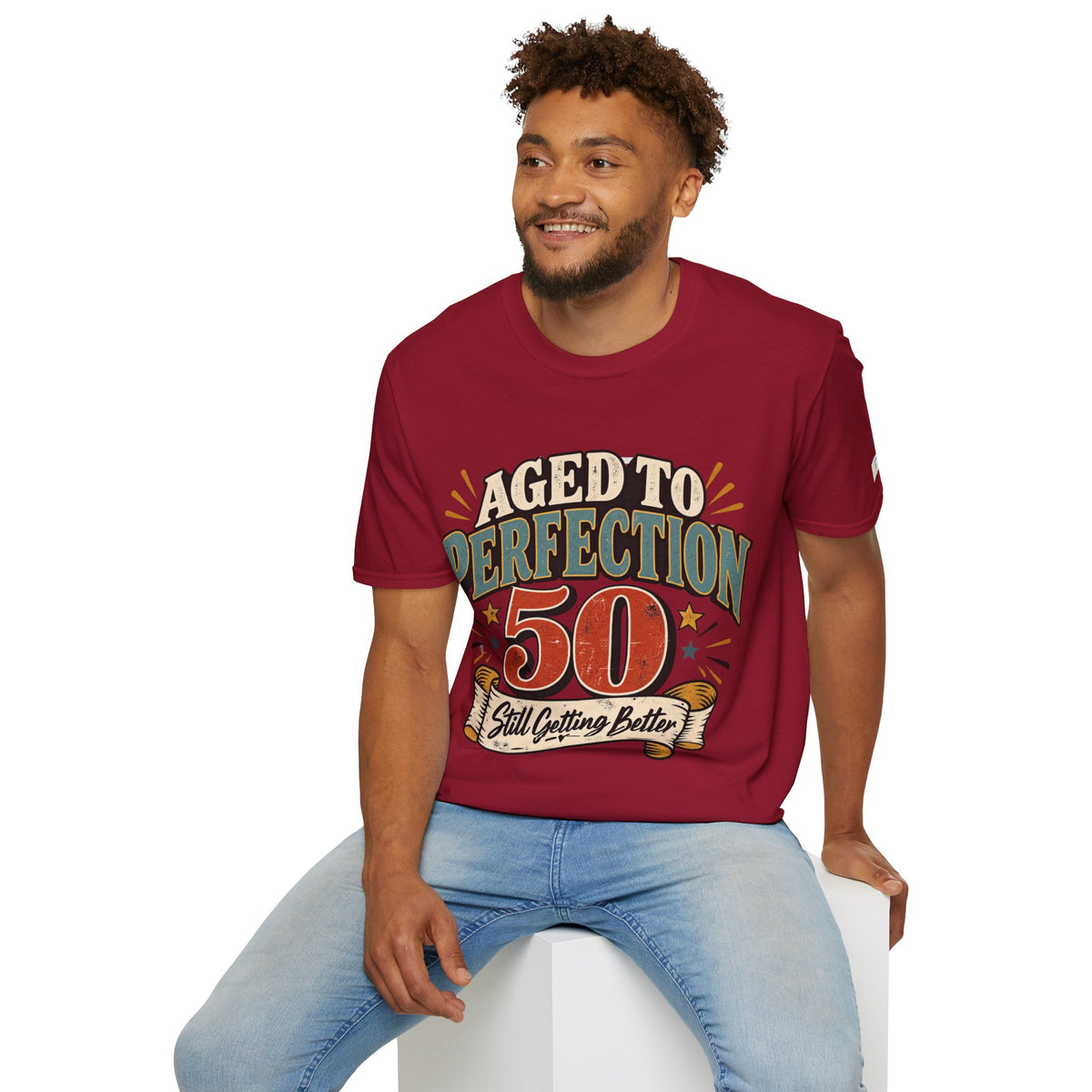 A man wearing a maroon short-sleeve graphic t-shirt with a retro-style 'Aged to Perfection 50' print, paired with light blue jeans, sitting on a white block against a white background.
