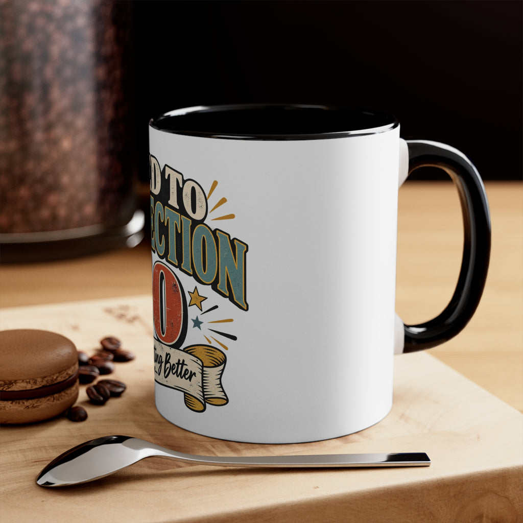 A white ceramic coffee mug with a black interior and handle, featuring a retro-style graphic that reads 'aged' with stars and a banner. The mug sits on a wooden surface next to a spoon, coffee beans, and a macaron, with a blurred coffee jar in the background.