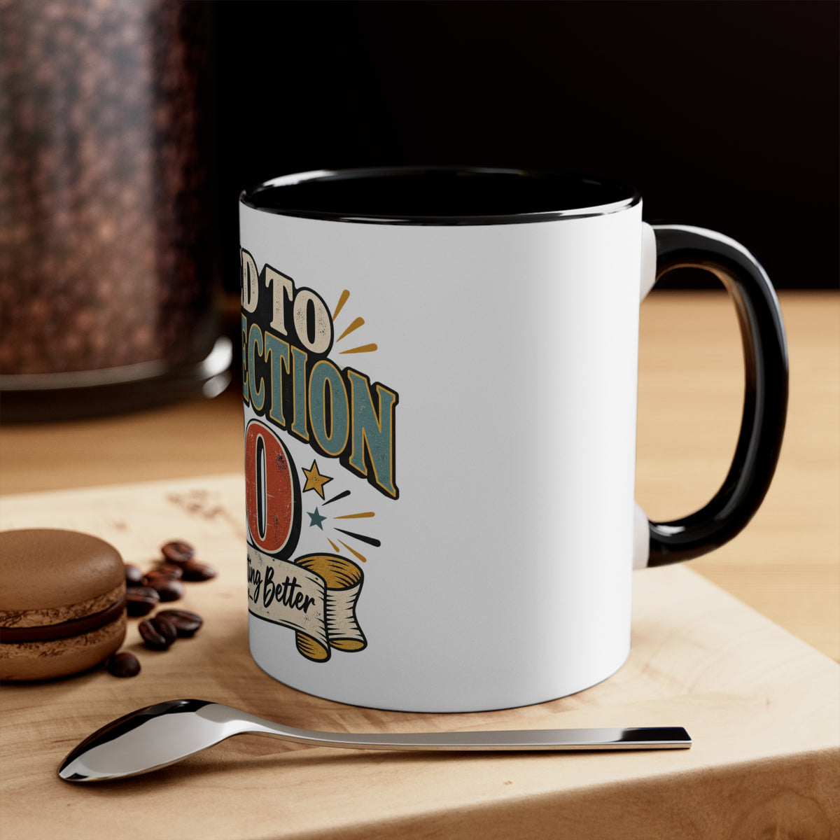 A white ceramic coffee mug with a black interior and handle, featuring a retro-style graphic that reads 'aged' with stars and a banner. The mug sits on a wooden surface next to a spoon, coffee beans, and a macaron, with a blurred coffee jar in the background.