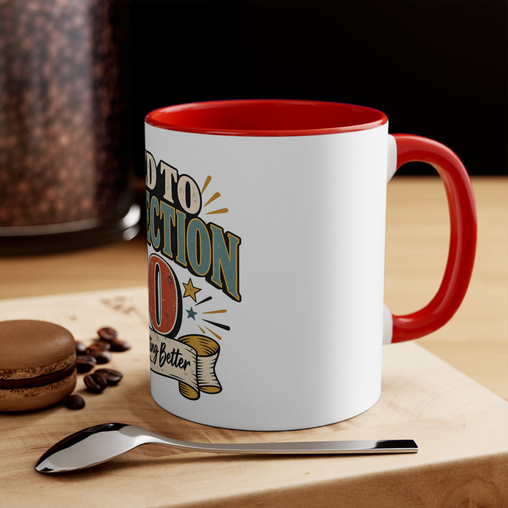 A white ceramic coffee mug with a red interior and handle, featuring a retro-style graphic that reads 'aged to perfection' with stars and a banner. The mug is styled on a wooden cutting board with a spoon, coffee beans, and a macaron in the background.
