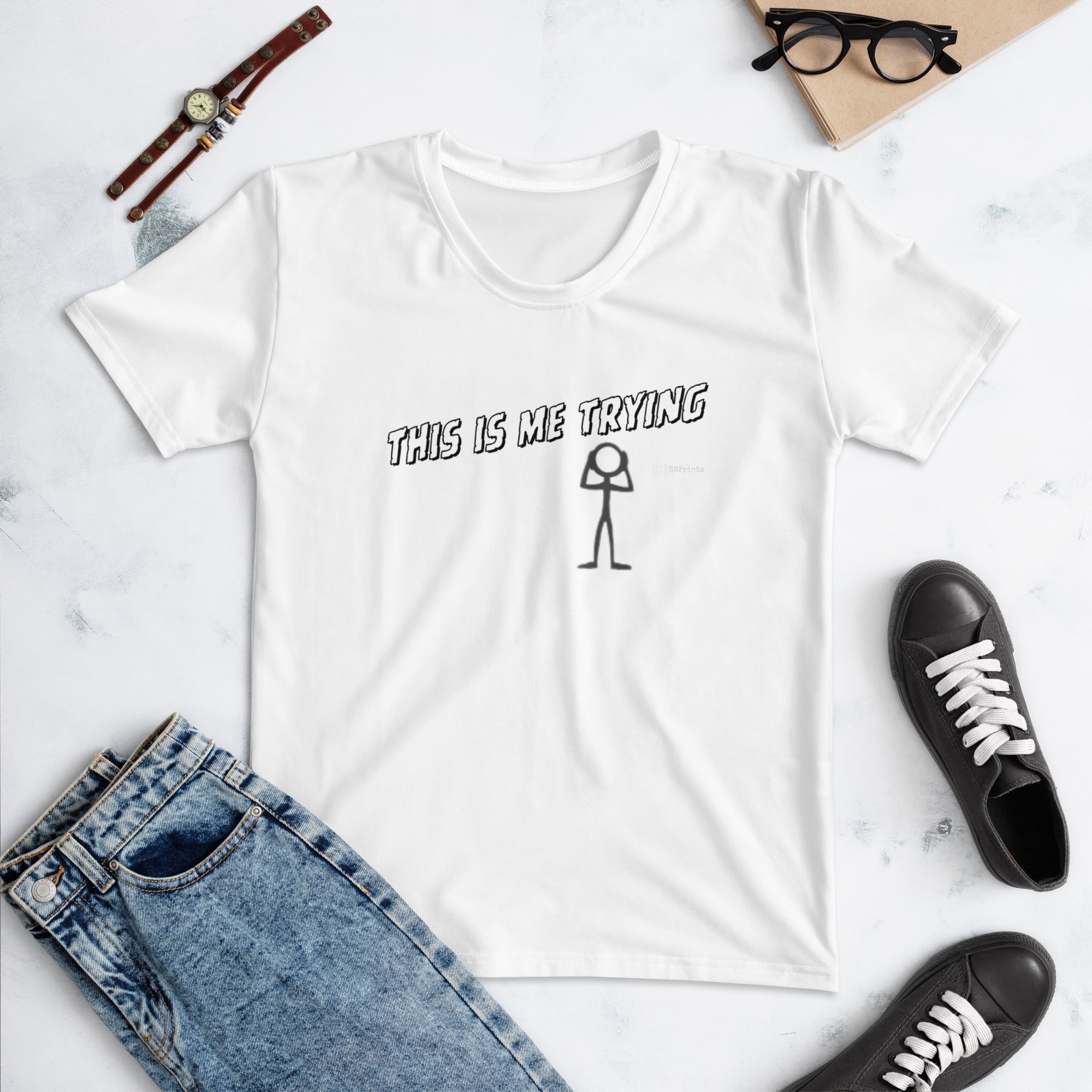 White t-shirt with 'This is me trying' text and stick figure graphic, surrounded by jeans, sneakers, and a watch on a light surface.