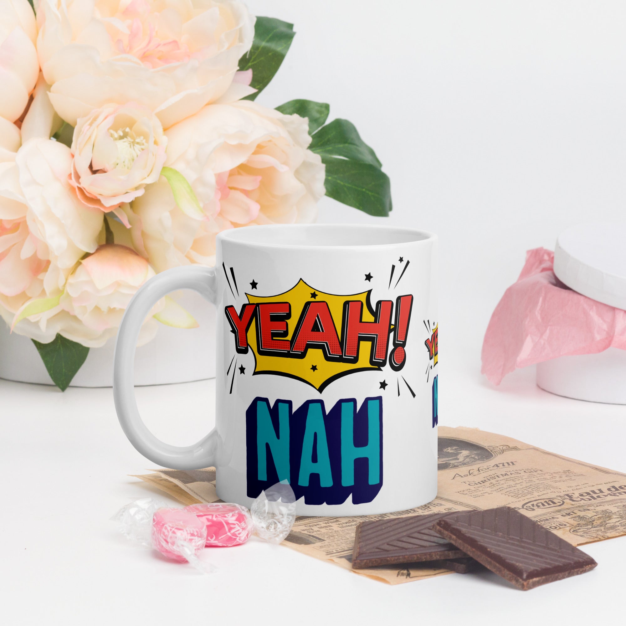 White mug with 'Yeah! Nah!' design on a table with flowers and snacks