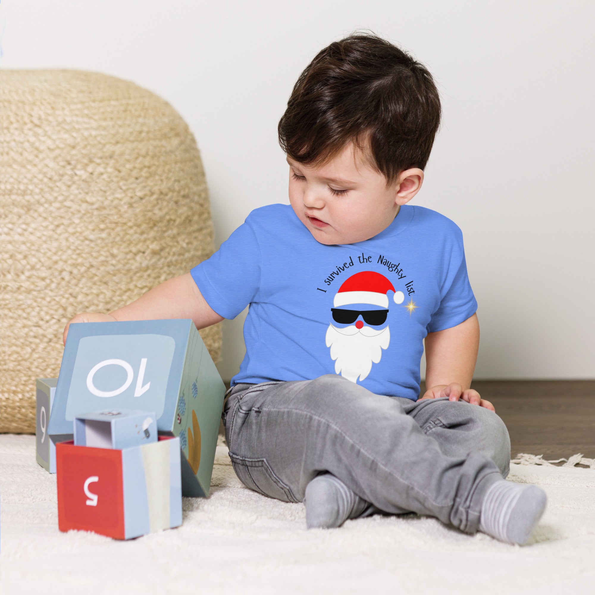 Child wearing a blue t-shirt with a graphic design of a Santa Claus wearing sunglasses, sitting on a carpeted floor.