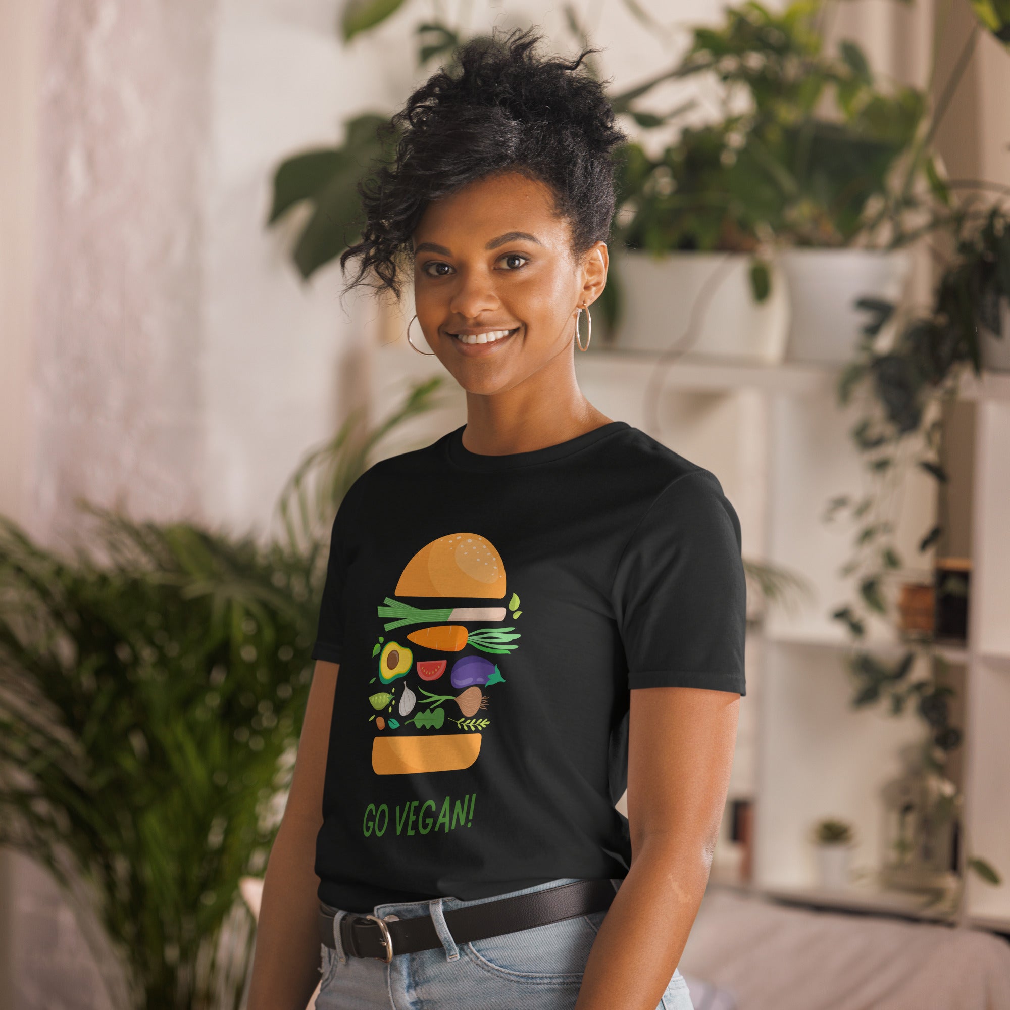 Person wearing a black t-shirt with a graphic of a burger and 'Go Vegan!' text, standing in a casual setting with plants.