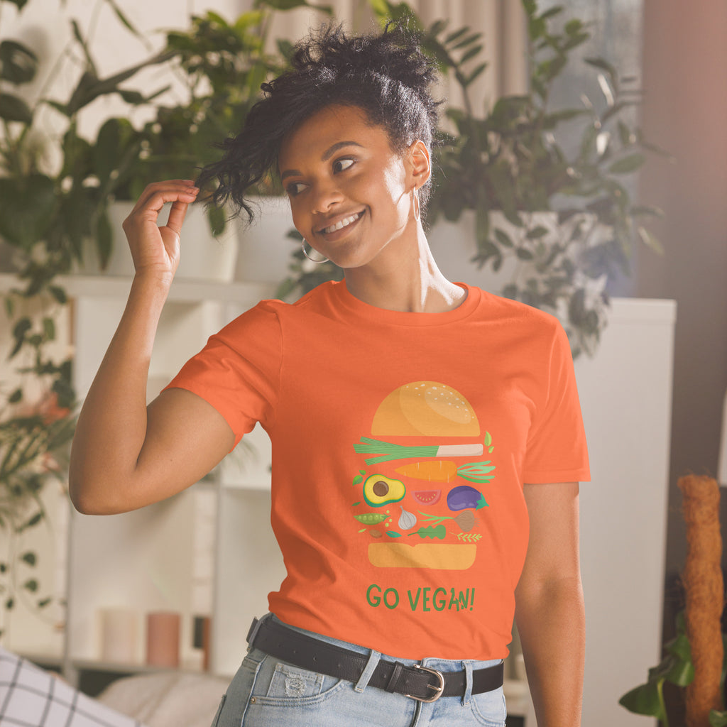 Person wearing an orange t-shirt with a vegan burger graphic and 'Go Vegan!' text, standing in a room with plants.