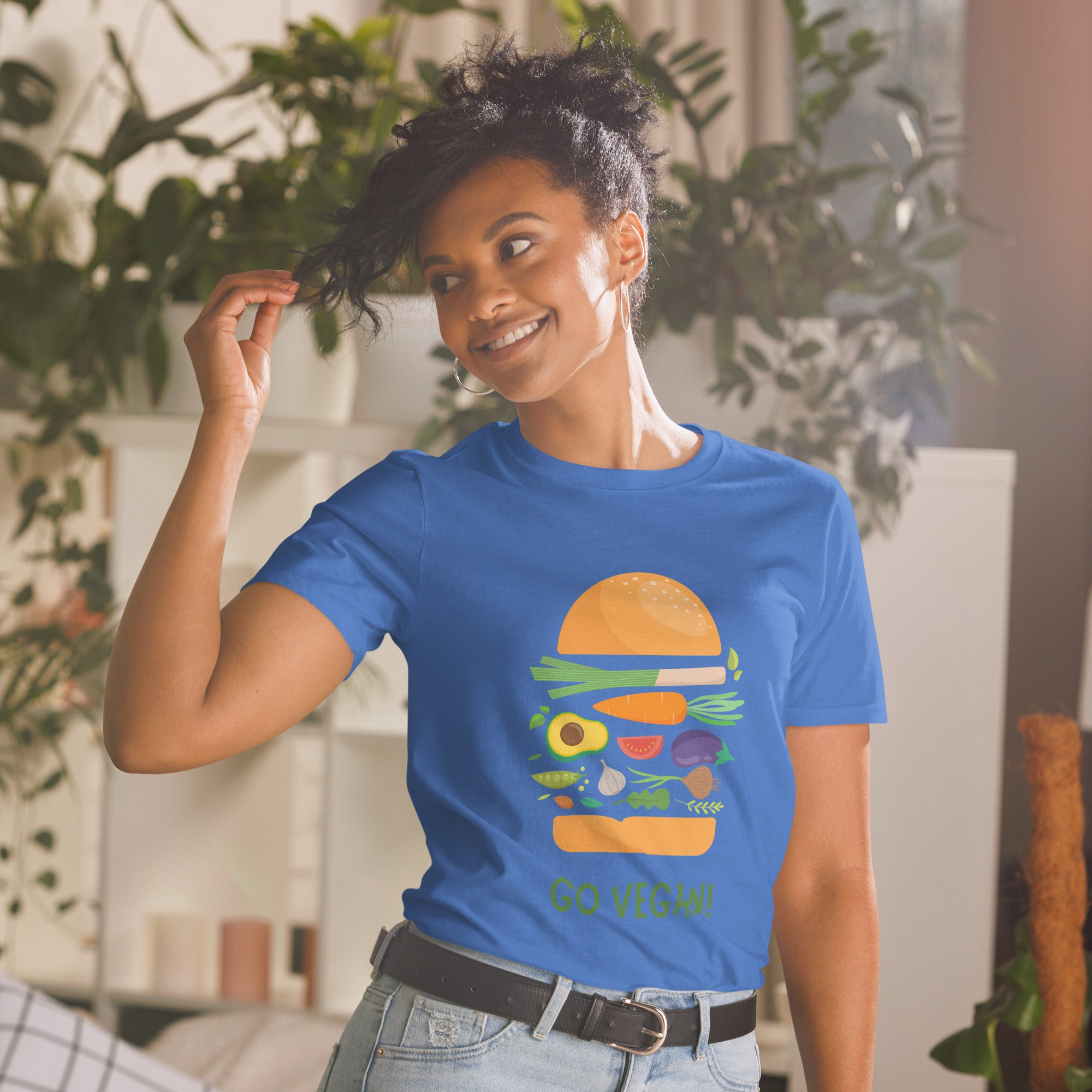 Woman wearing a blue t-shirt with a colorful graphic and 'Go Vegan!' text, standing in a room with plants.