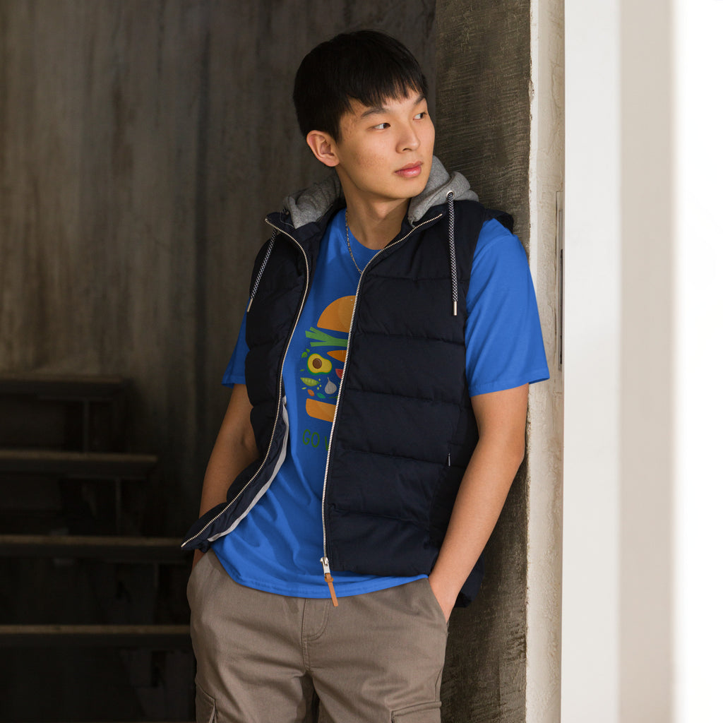 Person wearing a blue shirt with a colorful design and black vest leaning against a wall.