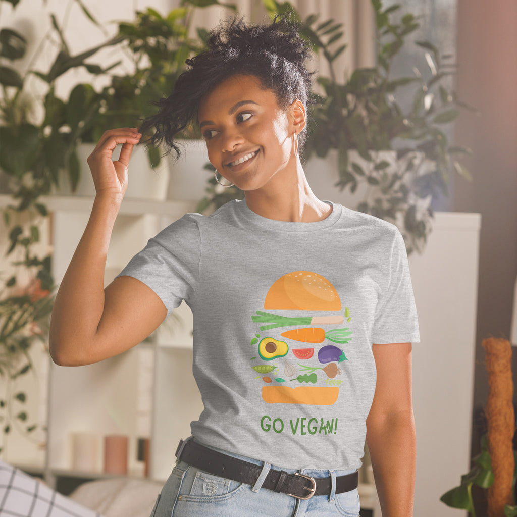 Woman wearing a gray t-shirt with a vegan burger graphic and 'Go Vegan!' text, standing in a room with plants.