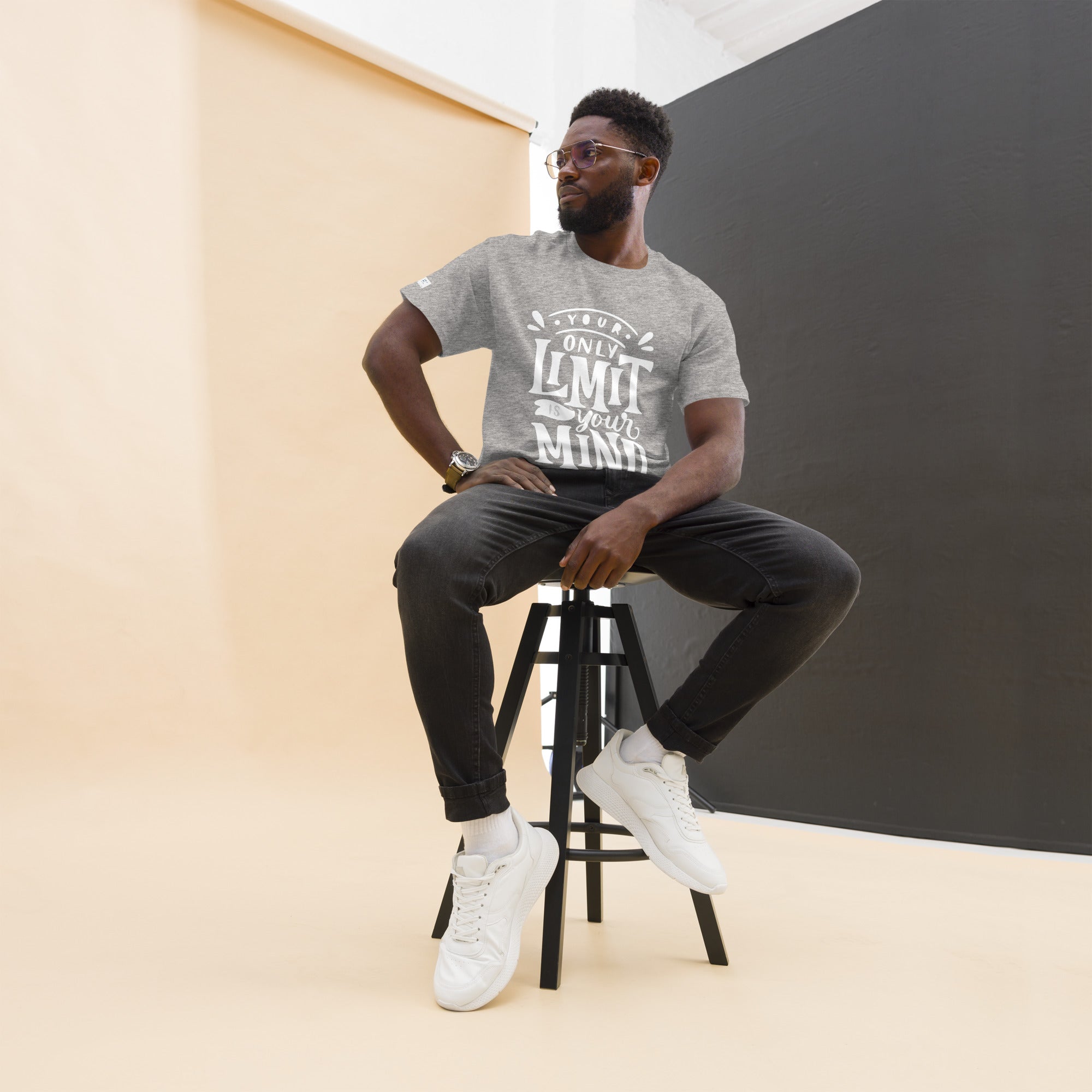 Man sitting on a stool wearing a gray t-shirt with text, black pants, and white sneakers.