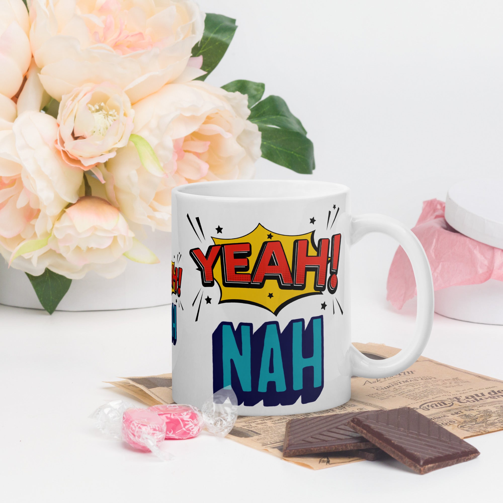 Mug with 'YEAH! NAH!' design on a table with flowers and candy