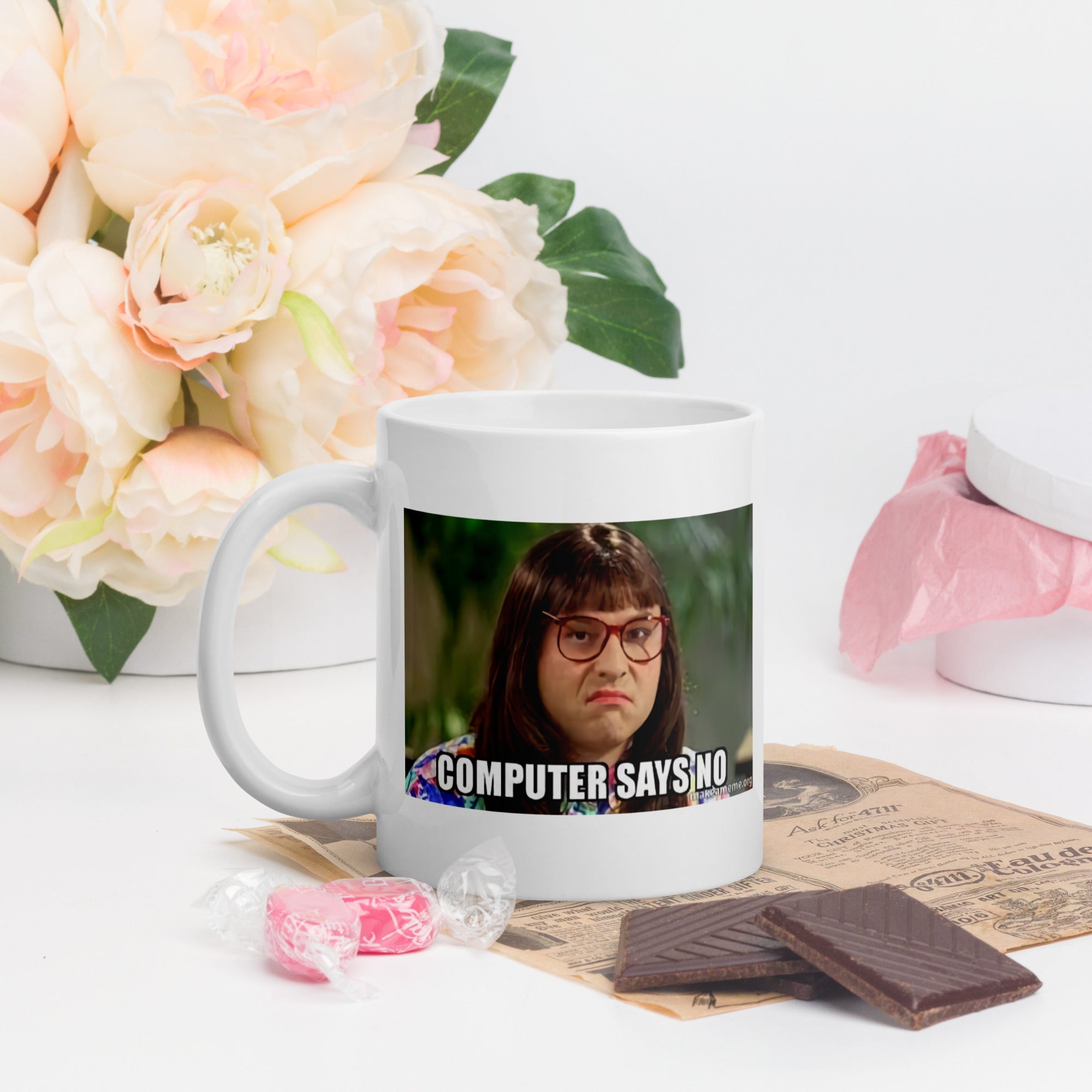 White mug with a printed design featuring a character and text 'COMPUTER SAYS NO' on a table with flowers and snacks.